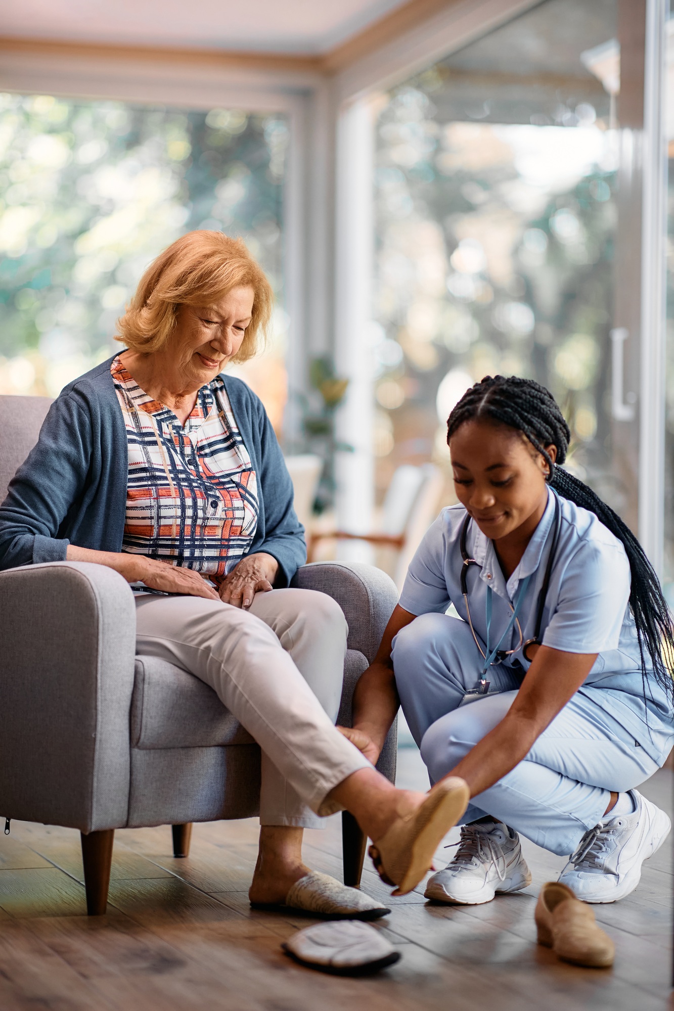 Young black nurse helping senior woman to take off her shoes in nursing home.